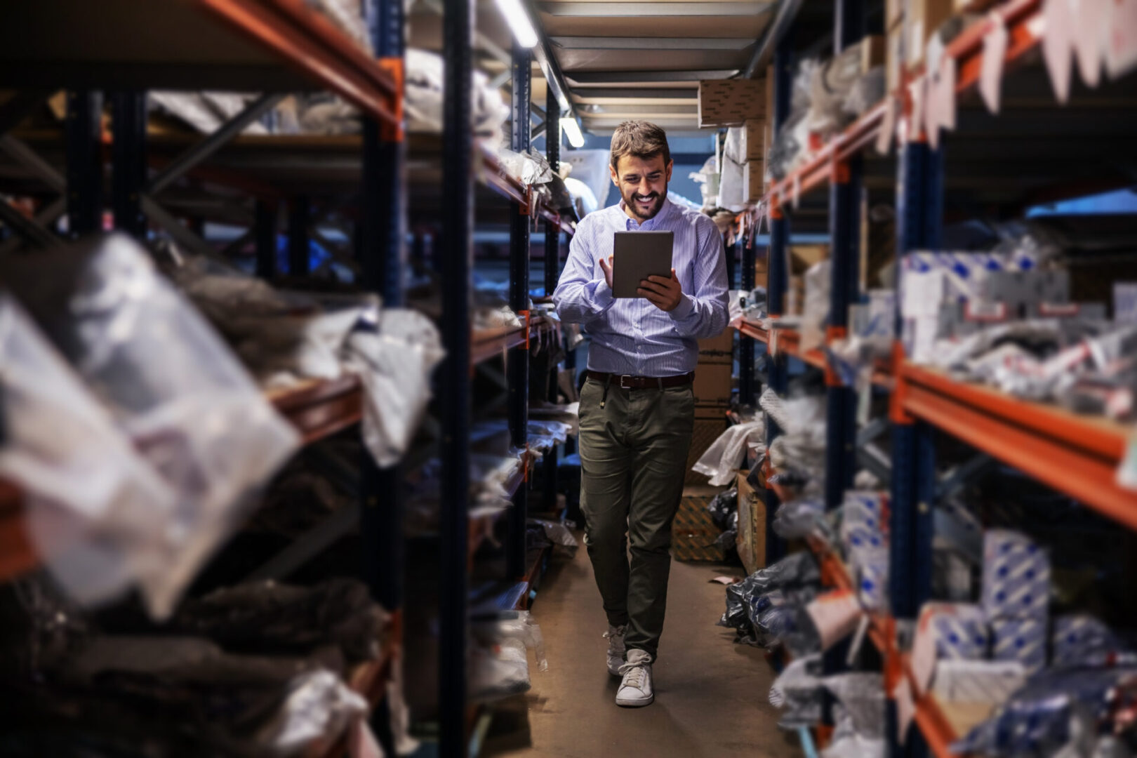 Full length of bearded supervisor walking trough storage of export firm, checking on goods and using tablet.